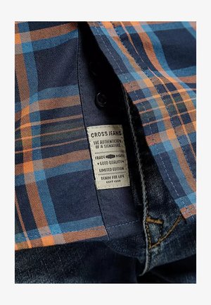 Niebieska i pomarańczowa koszula w kratę częściowo otwarta, odsłaniająca metkę materiałową z napisem "Cross Jeans" oraz pas dżinsów poniżej.