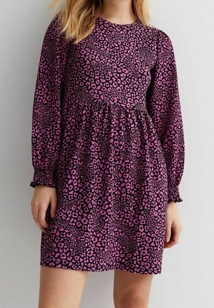 Woman wearing a purple and black leopard print long-sleeve mini dress with a gathered waist and elasticized cuffs, standing against a plain wall.