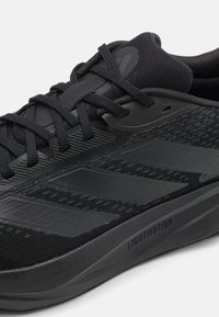 Gros plan d'une chaussure de sport noire avec une tige en mesh, des bandes latérales texturées, une semelle épaisse marquée Lightmotion, et des lacets noirs noués sur le dessus.
