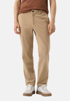Mann trägt beige Cordhose, weiße Sneaker mit braunen Sohlen und ein braunes Hemd, steht vor weißem Hintergrund.