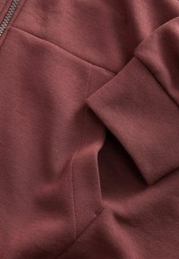 Bordeauxfarbenes Sweatshirtmaterial mit glatter Textur und sichtbaren Nähten, ausgestattet mit einem Reißverschluss und einer großen Vordertasche.