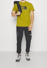 Olijfgroen t-shirt met zwart logo, zwarte broek met zijzakken en meerkleurige sneakers met grijze, oranje en zwarte accenten.
