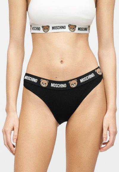 Γυναίκα που φοράει λευκό crop top και μαύρο μαγιό μπικίνι με το λογότυπο Moschino και εικόνες αρκουδάκια στις ελαστικές ζώνες.