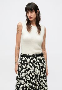 Ärmlös krämfärgad stickad topp kombinerad med en svart kjol med veck och ett vitt blommönster samt ett kontrasterande svart bälte.