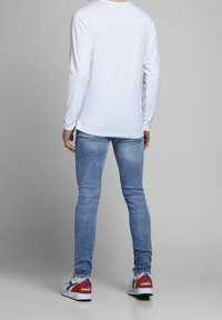 Persona vestida con una camisa blanca de manga larga, jeans ajustados de color azul claro y zapatillas blancas con acentos rojos y azules, de pie contra un fondo gris liso.