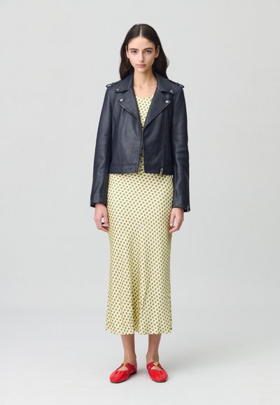 Veste en cuir noir de motard, manches courtes, robe jaune à pois bleus de longueur mi-longue, associée à des chaussures rouges avec un accent doré.