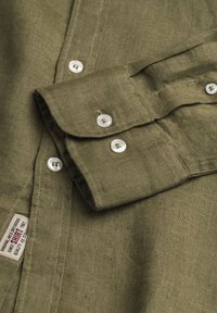 Camicia in lino verde oliva con polsini a pieghe, caratterizzata da bottoni in perla e un piccolo etichetta cucita sulla cucitura laterale. Il tessuto ha una trama visibile.