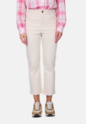 Femme portant un pantalon court de couleur crème, une chemise à carreaux rose rentrée à l’intérieur, et des baskets beige et blanches, debout devant un fond uni.