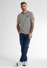 Proužkované bavlněné tričko v námořnické modři a bílé barvě, s kulatým výstřihem a malým logem na hrudi; zkombinováno s tmavě modrými džínami a bílými sportovními botami.