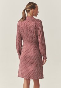 Rochie roșie cu mâneci lungi, cu un model geometric alb, o croială ușor conică și un guler. Material neted și lungime medie.