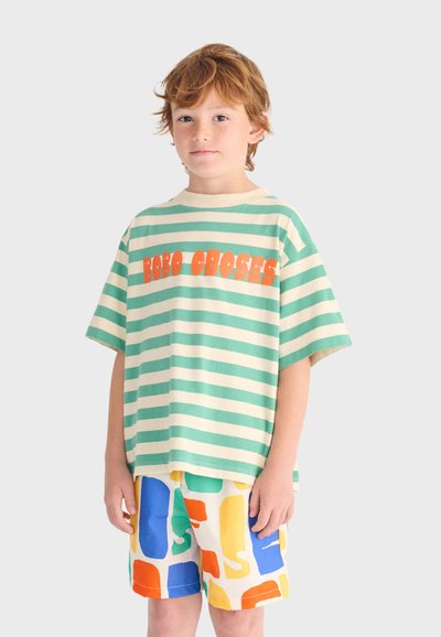 Junge trägt ein grün-cremefarben gestreiftes Hemd mit orangem Text und bunt gemusterte Shorts, steht vor einem hellen Hintergrund.