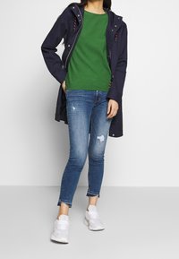Navy jacket with hood, green sweater, distressed blue jeans, and white sneakers. The jacket features a zip closure and drawstring accents.