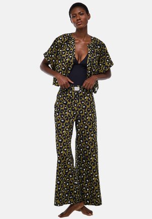 Femme portant une veste à manches courtes et un pantalon large assortis à imprimé léopard par-dessus un haut noir à décolleté en V profond, debout pieds nus sur un fond blanc.