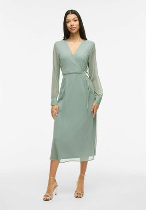 Grön, omlottdress med långa, genomskinliga ärmar och knytband i midjan. Slät textur, medellång, med en subtil lyster. Guldklackade sandaler.