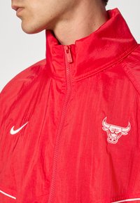 Rød vindjakke lavet af letvægtsmateriale, med høj krave, lynlås lukning, hvid Nike-logo og et Chicago Bulls emblem.