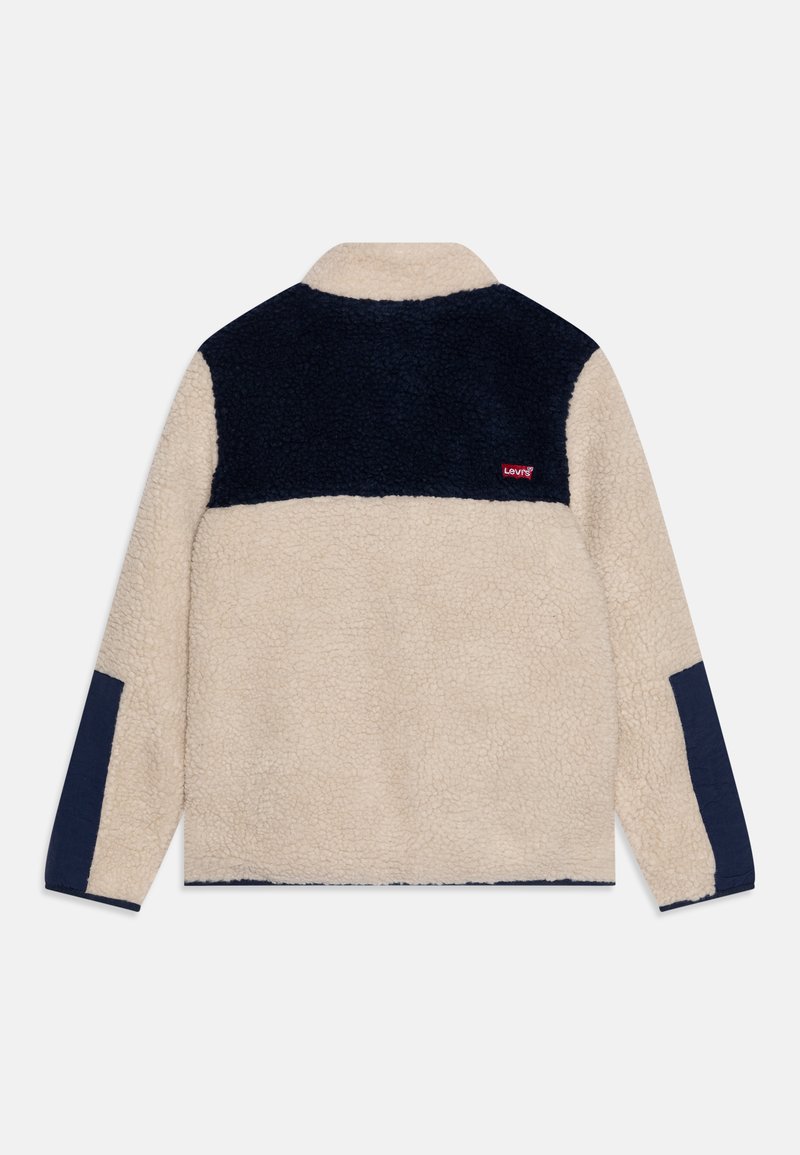 Chaqueta de forro polar beige y marino con mangas largas, yugo de hombro marino y parches en los codos, con un pequeño logo rojo de Levi's en el hombro derecho detrás.