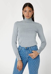 Maglione grigio chiaro con collo alto e maniche lunghe, decorato con bottoni laterali, abbinato a jeans a vita alta blu con un design a fantasia.