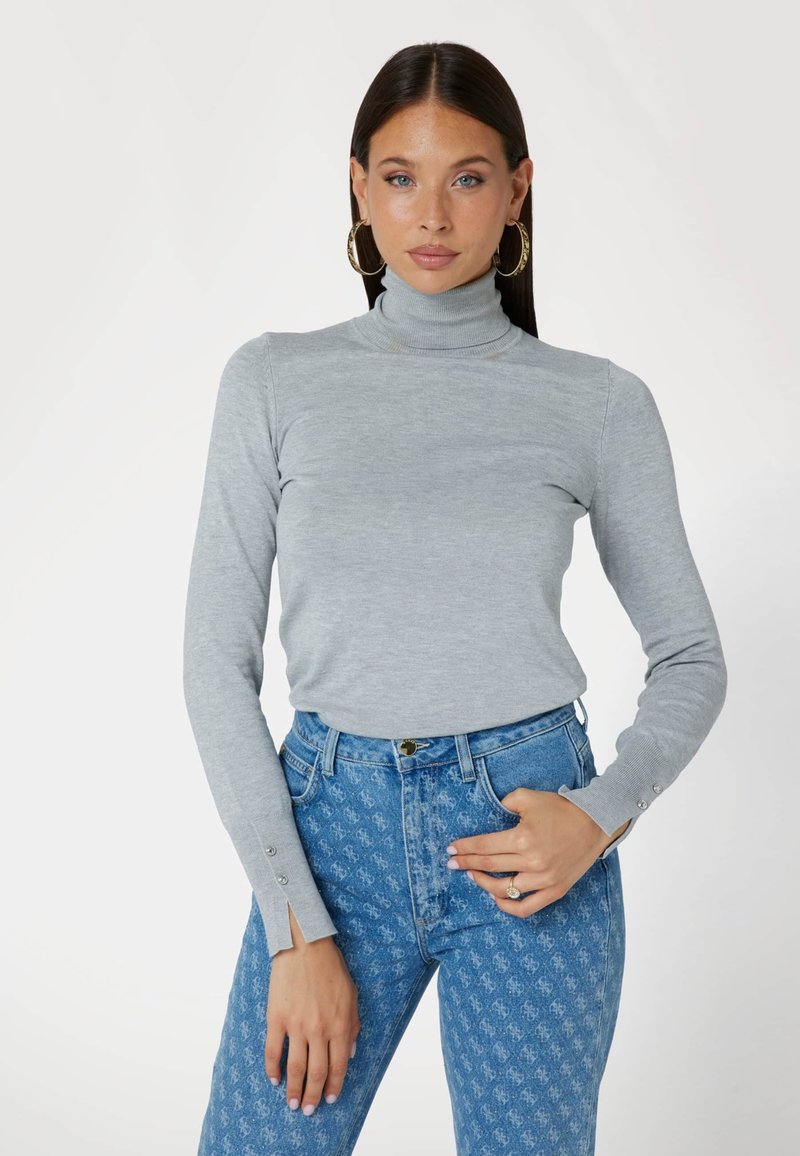 Maglione grigio chiaro con collo alto e maniche lunghe, decorato con bottoni laterali, abbinato a jeans a vita alta blu con un design a fantasia.