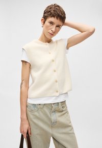 Κρεμ σαλιγκαρόμαυρο με κοντά μανίκια και στρογγυλή λαιμόκοψη, διακοσμημένο με κουμπιά στην μπροστινή πλευρά. Συνδυασμένο με λευκό μπλουζάκι και ανοιχτόχρωμα τζιν.