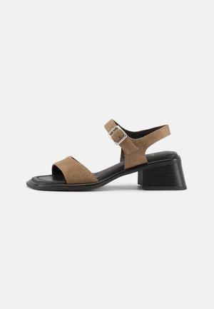 Smeđa sandala otvorenih prstiju od brušene kože s podesivim remenom na kopču i niskom crnom blok petom na bijeloj pozadini.