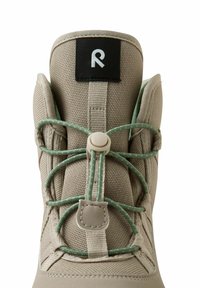 Bota textil beige con cuello acolchado, cordones elásticos verdes y una etiqueta negra con logo blanco en la parte superior.