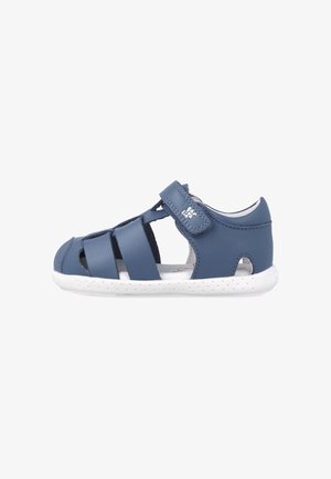 Blå lædersandal med elastiske stropper, åbne sider og en hvid, tekstureret gummisål. Har en velcro-lukning for nem justering.