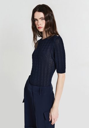 Frau mit langen braunen Haaren, die ein kurzärmliges, navyblaues Strickoberteil und passende navyblaue Hosen vor einem schlichten hellen Hintergrund trägt.