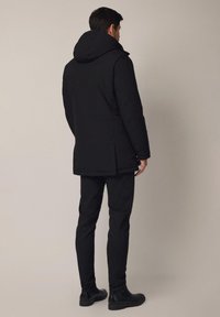 Czarna ocieplana parka z dużym kapturem, o prostym kroju, z bocznymi kieszeniami i teksturowanym materiałem dla zapewnienia ciepła. Nosi się ją na czarnych spodniach.