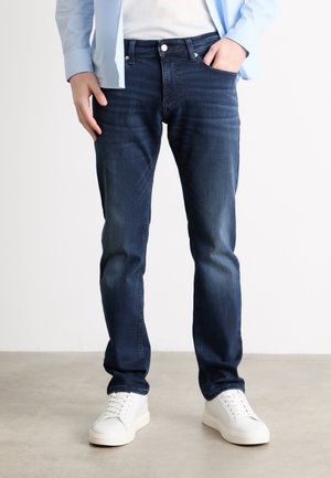 Hombre con vaqueros ajustados azul oscuro, zapatillas blancas, camiseta blanca y camisa de cuadros azul claro, de pie sobre un suelo gris.