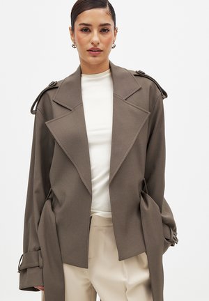 Mujer con el cabello peinado hacia atrás, vestida con una chaqueta oversize taupe sobre una camiseta blanca y pantalones beige, sobre un fondo blanco liso.