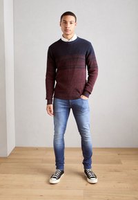 Jersey de punto a rayas en marrón y azul marino con cuello redondo, combinado con jeans ajustados azules y zapatillas negras sobre un suelo de madera.