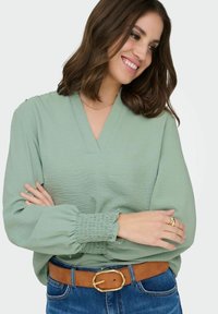 Femme aux cheveux bruns mi-longs portant un chemisier vert clair, une ceinture marron, un jean bleu, souriante et regardant sur le côté avec les bras croisés.