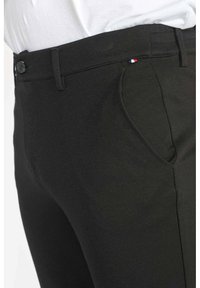 Pantalons noirs en tissu texturé avec une coupe ajustée, comportant une poche avant et un petit accent de drapeau tricolore près de la taille.