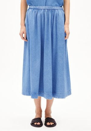 Femme portant une jupe midi bleue avec une ceinture élastique et des sandales noires à bout ouvert, debout sur un fond blanc.