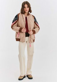 Prošívaná béžová bomber bunda s růžovými akcenty a černými vzorovanými rukávy, nošená přes texturovaný mahagonový svetr, spárovaná se smetanovými kalhotami.