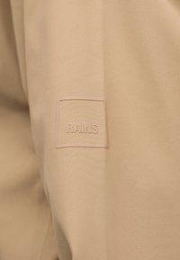 Μπεζ ύφασμα με ανάγλυφο τετράγωνο λογότυπο με τη λέξη "RAINS" στο κέντρο.