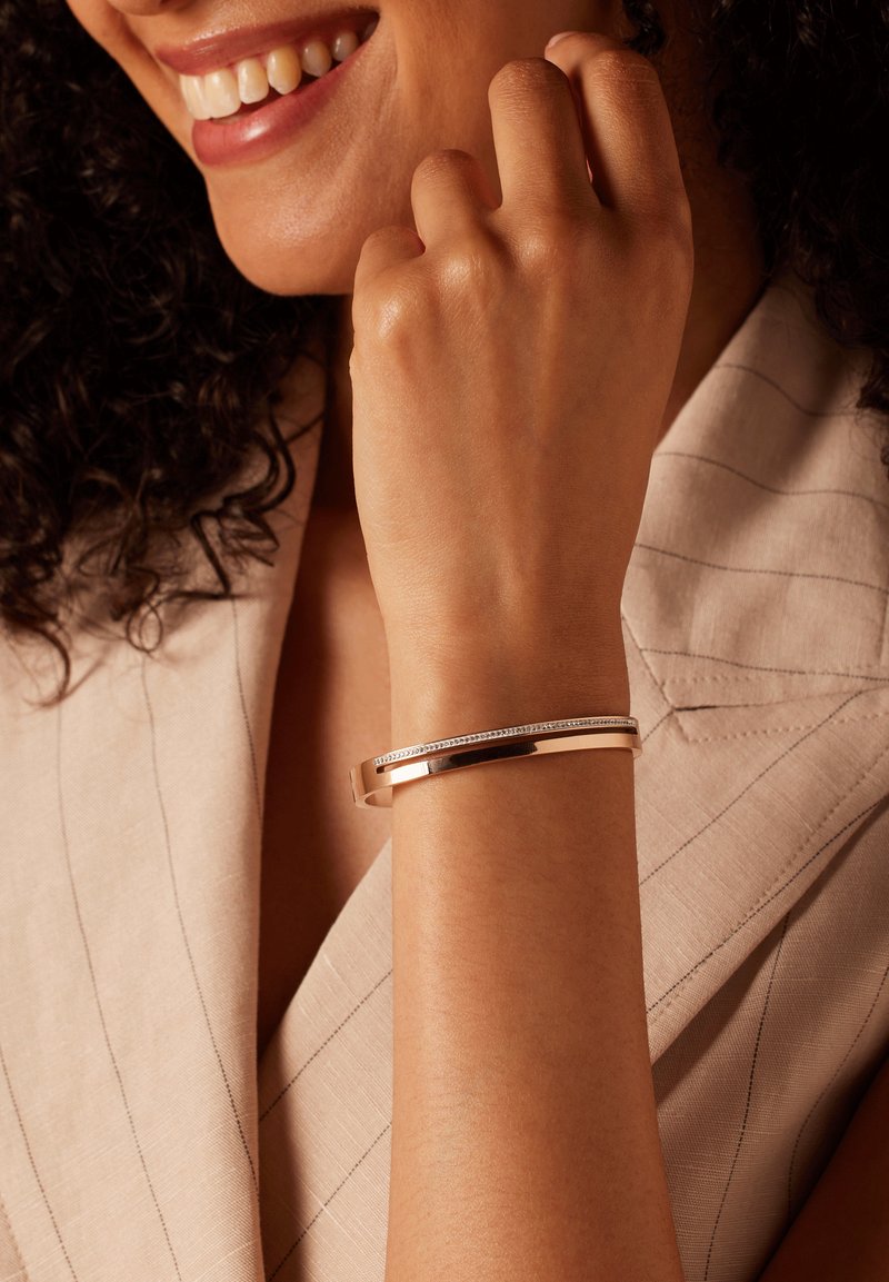 Metalen armband in goudkleur met een dunne band, voorzien van een rij kleine heldere stenen aan de bovenkant. Glad oppervlak, moderne design.