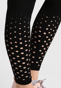 Detailní záběr na černé legíny s rovnoměrně rozmístěnými kruhovými výřezy, pod kterými je vidět pokožka, nošené s světle růžovými teniskami.