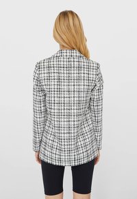 Woman with long blonde hair wearing a black-and-white checked blazer and black knee-length shorts, standing against a plain background.