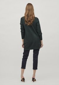 Lång, mörkgrön stickad tröja med ribbade muddar och nederkant, bärs över marinblå beskurna byxor. Modell visas från ryggen med löst, vågigt hår.