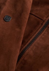 Materiale scamosciato marrone con una cerniera nera e un'etichetta logo in metallo. La vista ravvicinata mette in evidenza la texture e i dettagli di cucitura.