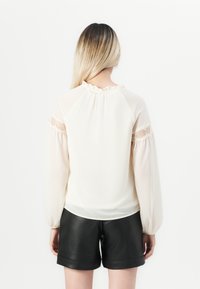 Blusa di colore crema chiaro con scollatura arricciata, maniche a palloncino trasparenti e dettagli in pizzo sulle maniche. Abbinata a pantaloni corti in pelle nera.
