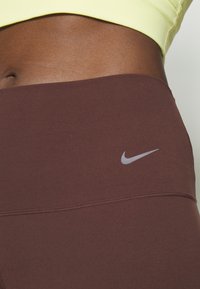 Legginsy Nike w ciemnobrązowym materiale, charakteryzujące się gładką teksturą i srebrnym logo w kształcie „swooshu”. Noszone z jasnosłoneczną bluzką.