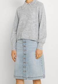 Femme portant un pull gris clair avec un col orné de perles et une jupe en jean bleu clair, mi-longue, avec des boutons à l'avant.
