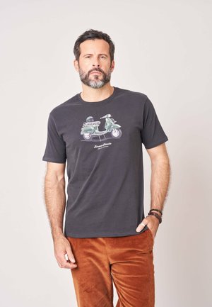 Man met baard die een zwart t-shirt draagt met een vintage scooterprint en bruine corduroy broek, staand met één hand in de zak.