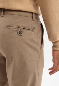 Brown trousers with a smooth texture, featuring a back pocket with a button closure and a branding tag on the pocket.