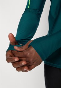 Αθλητικό μπλουζάκι με μακριά μανίκια σε τιρκουάζ από χρωση με εφαρμοστή γραμμή. Διαθέτει τρύπες για τον αντίχειρα στα μανίκια και αντίθετη νεονική λεπτομέρεια στον ώμο.