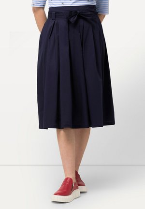 Kvinde iført navy plisseret nederdel med sløjfe i taljen, lyseblå stribet top og røde slip-on sneakers med hvide såler.