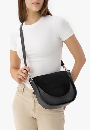 Kobieta ubrana w białą koszulkę i beżowe spodnie trzyma czarną skórzaną torebkę na ramię z zamszową przednią klapą.