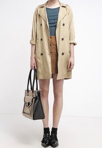 Manteau beige avec boutons double boutonnage, top empilé teal, jupe en suede marron, sac à main en vernis noir et mocassins noirs avec nœuds.